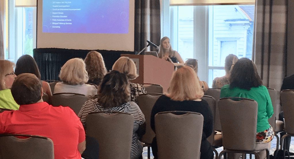 Shelby Wade (she/her) is facing an audience and gesturing while talking. She is standing in front of a podium with a projector screen and window behind her in a conference room. Shelby is a white woman with long blonde hair and she is wearing a dark top.
